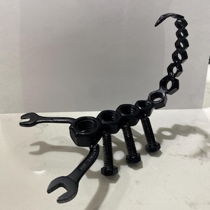 May include: Black metal scorpion sculpture crafted from wrenches, bolts, and nuts. The scorpion's body is formed from nuts and bolts, with wrenches used for the claws. The tail curves upwards, made from connected nuts.