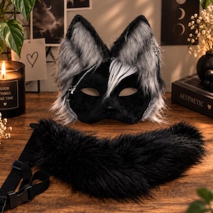 Op de afbeelding: Een zwart masker met hoge, puntige oren bedekt met lang, grijs en zwart imitatiebont, voorzien van een zilveren ketting. Een apart, dik zwart imitatiebont kraagje met een verstelbare nylon band en kunststof gesp ligt aan de voorzijde.