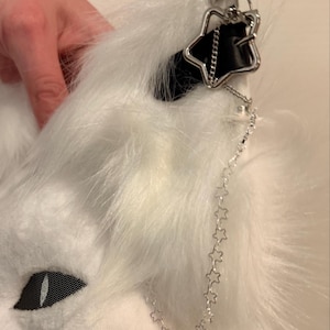 May include: A white faux fur animal ear headband featuring a black leather-like strap detail, a silver-toned star-shaped charm, and a delicate silver chain link strand composed of small connected star shapes hanging from the ear.