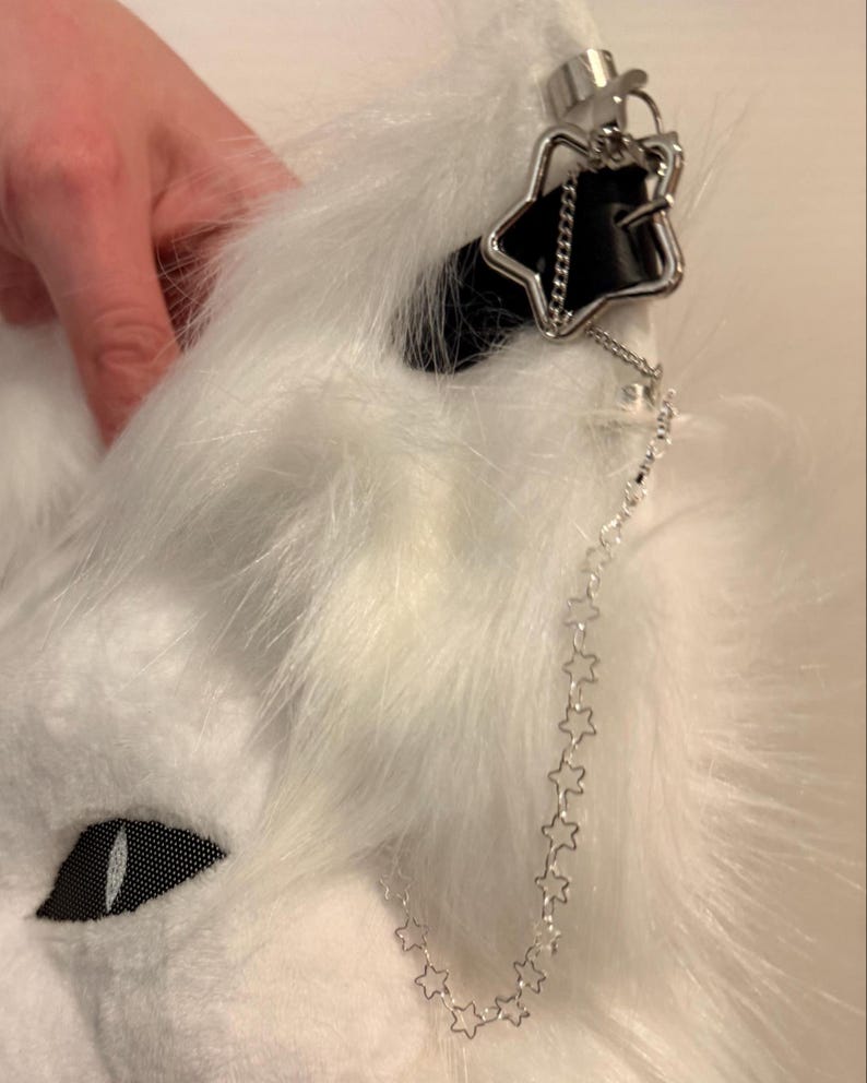 May include: A white faux fur animal ear headband featuring a black leather-like strap detail, a silver-toned star-shaped charm, and a delicate silver chain link strand composed of small connected star shapes hanging from the ear.