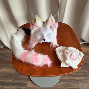 Può includere: Una maschera da volpe bianca con motivo floreale, orecchie in pelliccia sintetica rosa e bianca e fiocchi decorativi. Il set include una lunga coda soffice a righe bianche e rosa e un paio di guanti in peluche con cuscinetti rosa sui palmi.