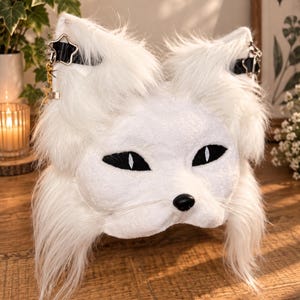 May include: A white faux fur animal mask featuring pointed ears with black straps and star-shaped metal charms. The mask has almond-shaped black eyes with white pupils, a small black nose, and long white whiskers extending from the muzzle area.
