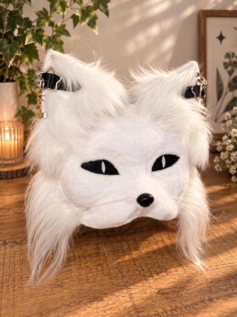 May include: A white faux fur animal mask featuring pointed ears with black straps and star-shaped metal charms. The mask has almond-shaped black eyes with white pupils, a small black nose, and long white whiskers extending from the muzzle area.