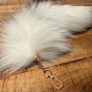 May include: A long, fluffy white faux fur tail accessory featuring a gold-toned metal cap at the base. A short gold chain connects the tail to a gold-toned lobster claw clasp, designed for easy attachment to bags, keys, or other personal items.
