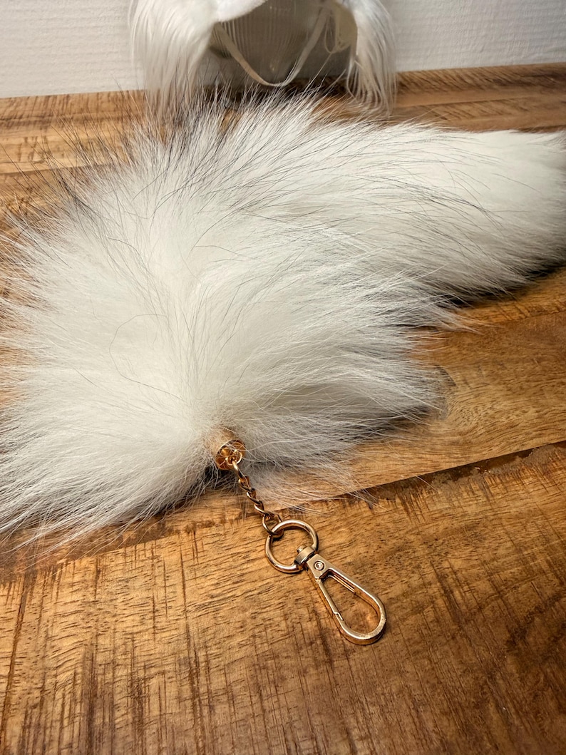 May include: A long, fluffy white faux fur tail accessory featuring a gold-toned metal cap at the base. A short gold chain connects the tail to a gold-toned lobster claw clasp, designed for easy attachment to bags, keys, or other personal items.
