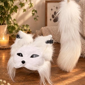 May include: A white faux fur animal mask featuring pointed ears with black star-shaped buckles, almond-shaped black eyes, and a small black nose, accompanied by a long, matching white faux fur tail attached to a gold-toned metal keychain ring.