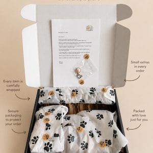 May include: An open black cardboard shipping box containing items wrapped in white tissue paper with a black paw print pattern. A thank you note from Flowykin and a small clear plastic bag with a metal charm are placed on top of the tissue paper.