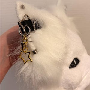 May include: A white faux fur animal ear headband featuring a black strap and silver-toned metal hardware. The ear is decorated with a silver star charm, a small silver hoop, a dangling gold-toned star charm, and a silver-toned spike pendant.