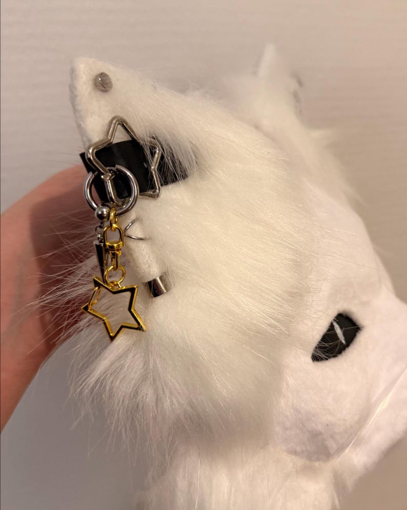 May include: A white faux fur animal ear headband featuring a black strap and silver-toned metal hardware. The ear is decorated with a silver star charm, a small silver hoop, a dangling gold-toned star charm, and a silver-toned spike pendant.