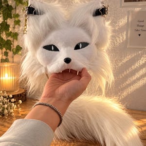 May include: A white faux fur animal mask featuring pointed ears, black almond-shaped eyes, and a small black nose. The mask is adorned with black straps, silver star-shaped charms, and dangling gold-toned chains. A long, matching white faux fur tail is attached.