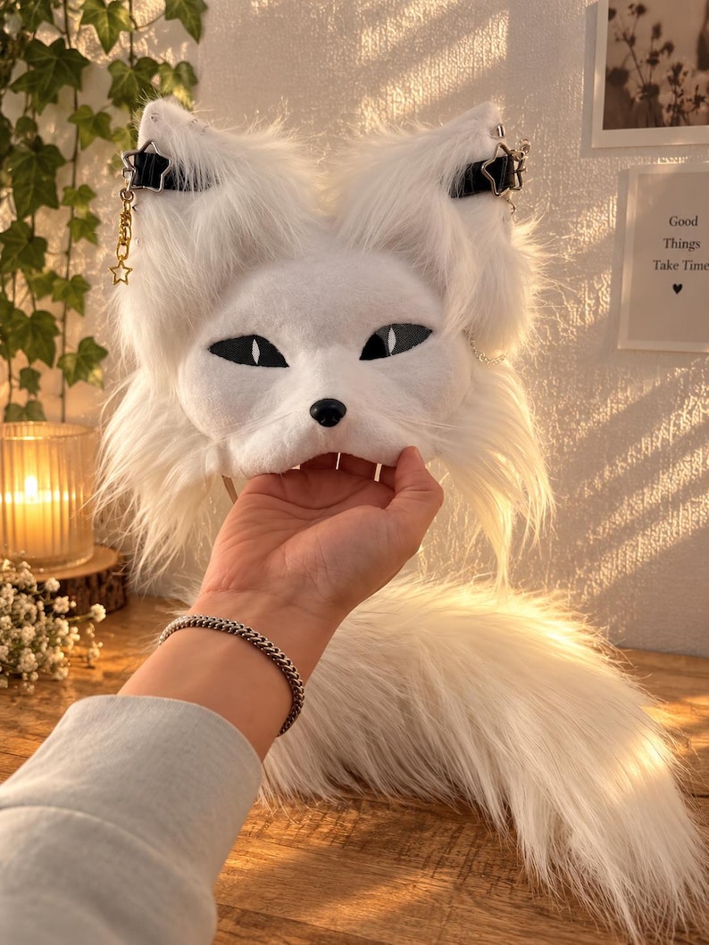 May include: A white faux fur animal mask featuring pointed ears, black almond-shaped eyes, and a small black nose. The mask is adorned with black straps, silver star-shaped charms, and dangling gold-toned chains. A long, matching white faux fur tail is attached.