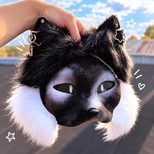 Op de afbeelding: Een zwart-wit dierenmasker van imitatiebont met puntige oren voorzien van stervormige metalen bedels, gaas over de ogen en een kleine zwarte neus. Het masker heeft lang wit bont aan de zijkanten en een fijn zilverkleurig kettinkje over het voorhoofd.