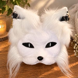 May include: A white faux fur animal mask featuring pointed ears, black almond-shaped eyes with white pupils, a small black nose, and long white whiskers. The mask includes black straps across the ears adorned with silver star-shaped charms.