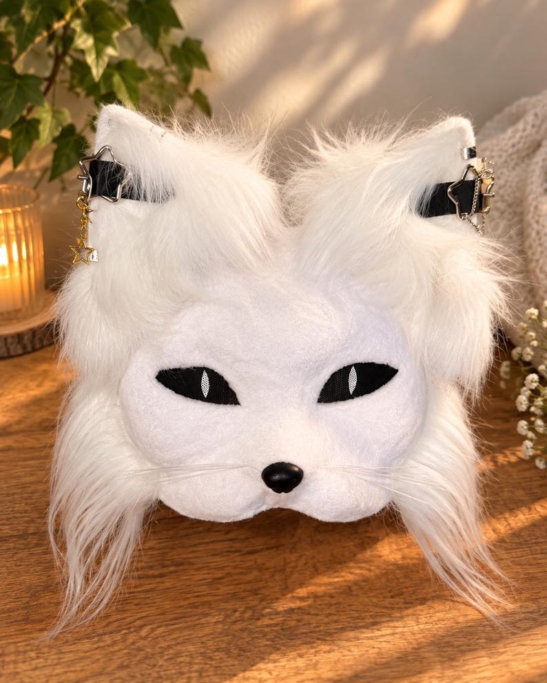 May include: A white faux fur animal mask featuring pointed ears, black almond-shaped eyes with white pupils, a small black nose, and long white whiskers. The mask includes black straps across the ears adorned with silver star-shaped charms.