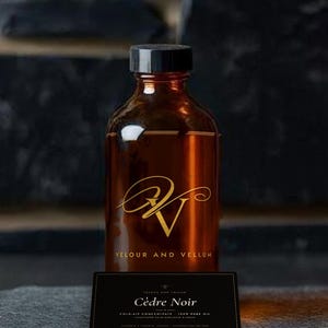 Peut inclure: Une bouteille en verre ambré avec un bouchon noir, arborant un logo doré et le texte VELOUR AND VELLUM. Devant se trouve une étiquette rectangulaire noire avec le texte Cèdre Noir, COLD-AIR CONCENTRATE, 100% PURE OIL et ASSEMBLED IN TORONTO, CANADA.