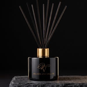 May include: A black glass reed diffuser bottle with a gold-coloured collar and a set of thin black reeds. The front of the bottle features a gold logo with the text VELOUR AND VELLUM printed in a serif font below the stylised initials.