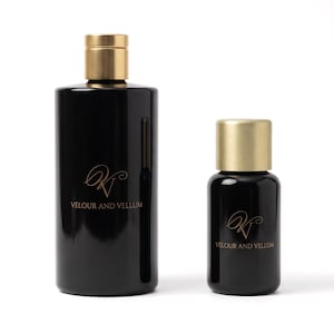May include: Two cylindrical black glass bottles with brushed gold caps. The larger bottle and the smaller bottle both feature a gold logo with the text VELOUR AND VELLUM printed on the front surface of the glossy black glass containers.