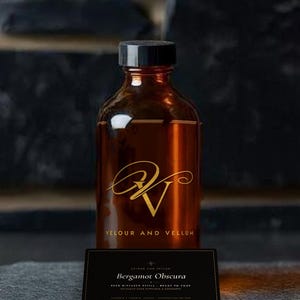Puede incluir: Una botella de vidrio ámbar con tapa negra, que presenta un logotipo dorado y el texto VELOUR AND VELLUM. Al frente se encuentra una etiqueta rectangular negra con el texto Bergamot Obscura, REED DIFFUSER REFILL - READY TO POUR y ASSEMBLED IN TORONTO.