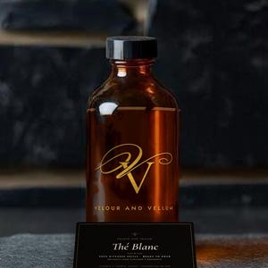 May include: An amber glass bottle with a black cap featuring a gold logo and the text VELOUR AND VELLUM. In front sits a black rectangular label with the text Thé Blanc, REED DIFFUSER REFILL, READY TO POUR, RECHARGE POUR DIFFUSEUR À BÂTONNETS.