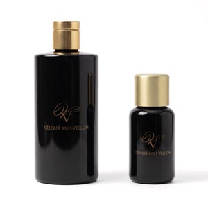May include: Two cylindrical black glass bottles with brushed gold caps. The larger bottle and the smaller bottle both feature a gold logo with the text VELOUR AND VELLUM printed on the front surface of the glossy black glass containers.