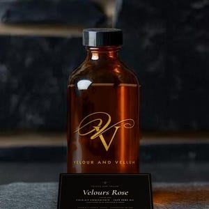 Peut inclure: Une bouteille en verre ambré avec un bouchon noir, arborant un logo doré et le texte VELOUR AND VELLUM. Devant se trouve une étiquette rectangulaire noire avec le texte Velours Rose, COLD-AIR CONCENTRATE, 100% PURE OIL, ASSEMBLED IN TORONTO, CANADA.