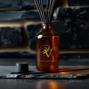 May include: An amber glass reed diffuser bottle with black reeds inserted. The bottle features a gold logo with the text "Velour and Vellum" printed on the front. A separate black cylindrical cap sits to the left of the glass bottle.