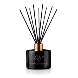 May include: A black glass reed diffuser bottle with a gold-coloured collar and ten thin black reeds. The front of the bottle features a gold-coloured logo with the text VELOUR AND VELLUM printed in a serif font below the stylised initials.