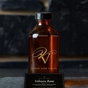 May include: An amber glass bottle with a black cap featuring a gold logo and the text Velour and Vellum. In front sits a black rectangular label with the text Velours Rose, Reed Diffuser Refill, Ready to Pour, and other small printed details.