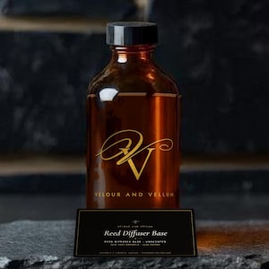 Puede incluir: Una botella de vidrio ámbar con tapa de rosca negra, con un logotipo dorado y el texto VELOUR AND VELLUM. Frente a ella hay una etiqueta rectangular negra con el texto Reed Diffuser Base, Velour and Vellum y Unscented en letras blancas.
