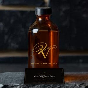 May include: An amber glass bottle with a black screw cap, featuring a gold logo and the text VELOUR AND VELLUM. In front sits a black rectangular label with the text Reed Diffuser Base, Velour and Vellum, and Unscented in white lettering.
