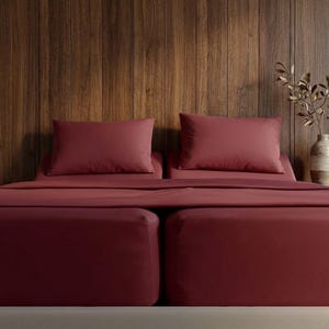 May include: A bed with burgundy-colored bedding, including two pillows and a fitted sheet. The bed is against a wooden wall. The color of the bedding is consistent throughout, creating a cohesive and inviting look. The scene is well-lit, highlighting the texture of the fabric.