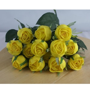 May include: A bunch of yellow artificial roses with green leaves. The roses are fully open, with tightly packed petals. The arrangement is on a light-coloured wooden surface, creating a vibrant display.