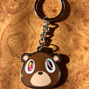 Könnte beinhalten: Ein brauner Bären-Schlüsselanhänger mit einer silbernen Kette und einem Ring. Der Bär hat große, farbenfrohe Augen mit rosa, gelben und blauen Akzenten. Der Schlüsselanhänger ist aus Metall und hat ein glänzendes Finish.