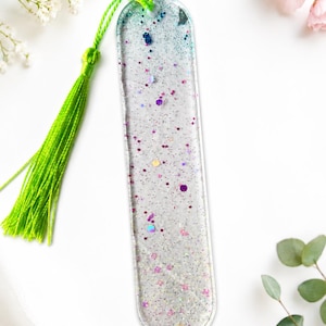 May include: A clear resin bookmark with embedded glitter in shades of purple, blue, and silver. It has a bright green tassel attached at the top. The bookmark is rectangular with rounded edges.