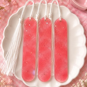 May include: Three pink resin bookmarks with white tassels, displayed on a white scalloped plate. The bookmarks have a glittery, speckled appearance. The background includes pink flowers and a pink heart-shaped dish.