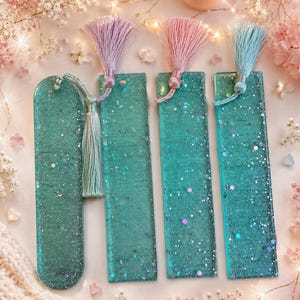 May include: Four teal resin bookmarks with glitter and tassels. One bookmark is curved at the top, while the other three are rectangular. Each bookmark has a different colored tassel: lavender, pink, and teal. The bookmarks are on a light pink surface with flowers.