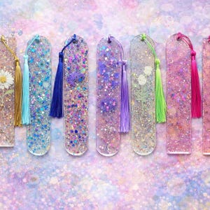 May include: A collection of handmade resin bookmarks with glitter and floral accents. Each bookmark features a unique color scheme, including gold, blue, purple, and pink, and is adorned with a matching tassel. The bookmarks are approximately 15 cm in length.