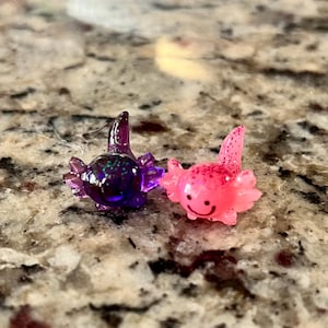 Op de afbeelding: Twee kleine, kleurrijke axolotl-beeldjes. Eén is paars met glitter en de andere is roze met glitter en een lachend gezicht. De beeldjes staan op een gespikkelde ondergrond.