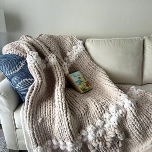May include: A chunky knit throw blanket in a soft beige color featuring thick, plush yarn loops. The blanket is accented with horizontal stripes of white, textured bobble-style yarn clusters and is draped over a sofa with a book resting on top.