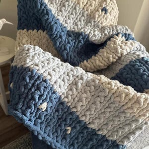 May include: A chunky knit blanket in shades of blue, cream, and gray. The blanket features thick, textured yarn in a striped pattern. The cozy throw is draped over a chair, showcasing its soft, inviting appearance. The blanket is a perfect addition to any home decor.