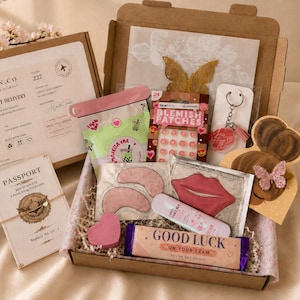 May include: An exam care package gift box containing a variety of items including a passport-style booklet, blemish patches, under-eye masks, a lip mask, a keychain, and a candy bar labelled Good Luck On Your Exam, all inside a cardboard box.