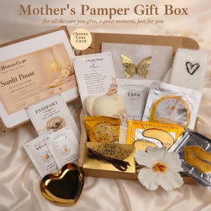 May include: A Mother's Pamper Gift Box containing self-care products. The box includes a gold heart-shaped dish, eye masks, bath salts, a butterfly decoration, and a white flower. The box is presented on a satin surface.