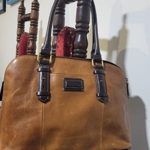 May include: A brown leather handbag with dark brown handles and accents. The bag has a rectangular shape with a curved top and a small rectangular label on the front. The bag is hanging on a wooden chair.
