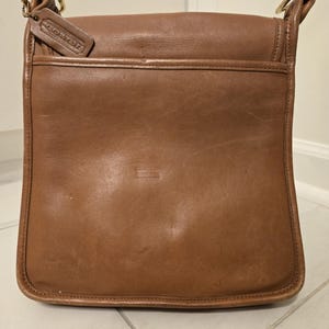 Könnte beinhalten: Braune Coach-Umhängetasche aus Leder mit Klappenverschluss und Schulterriemen. Die Tasche hat ein kleines Lederetikett mit dem Coach-Logo und goldfarbene Hardware. Die Tasche ist rechteckig und hat einen klassischen Vintage-Stil.