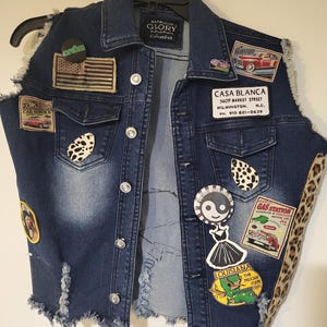 Könnte beinhalten: Dunkelblaue Jeansweste mit verschiedenen Aufnähern, darunter eine amerikanische Flagge und Motive für den Autoservice. Die Weste hat einen Used-Look mit ausgefransten Kanten und einem Leopardenmuster-Akzent an der Seite.