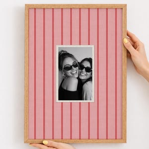 May include: A wooden framed picture with a pink and red striped background. The picture inside the frame is a black and white photo of two smiling women wearing sunglasses. The frame is being held by two hands.