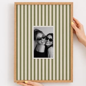 Pode incluir: Uma moldura de madeira clara com uma fotografia de duas pessoas sorrindo usando óculos de sol. A moldura tem um fundo listrado verde e branco. A foto está centralizada e é segurada por duas mãos.