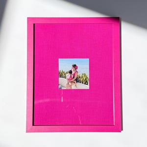 May include: A rectangular picture frame with a vibrant hot pink fabric-textured matting. Centered within the frame is a square photograph depicting two individuals sitting together outdoors near a body of water with green foliage in the background.
