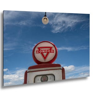 May include: A vintage Conoco gas pump with a red and white sign against a blue sky. The sign features the Conoco logo, a red triangle with the brand name. A light hangs above the pump.