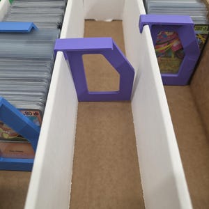 May include: Card storage box with dividers. The box has white cardboard dividers and purple plastic card holders. The cards are in protective sleeves. The box is brown on the inside.